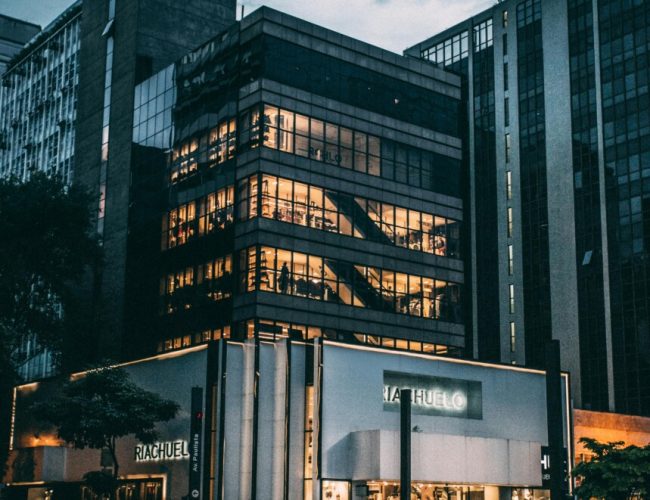Night view of a city block with illuminated store, modern architecture, and cloudy sky.